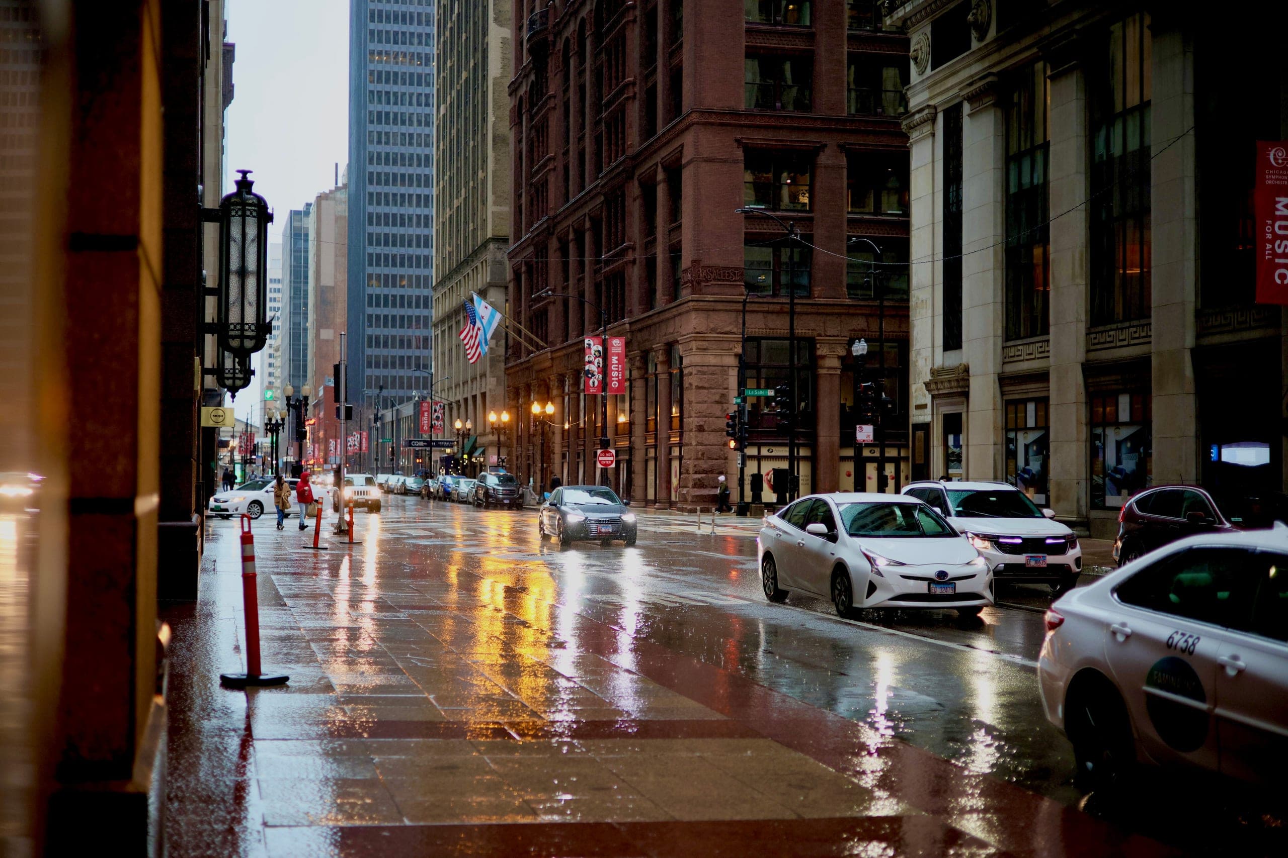 A city street at dusk after rain, wet asphalt reflecting street lamps and lit shopfronts, parked cars receding toward tall stone buildings.