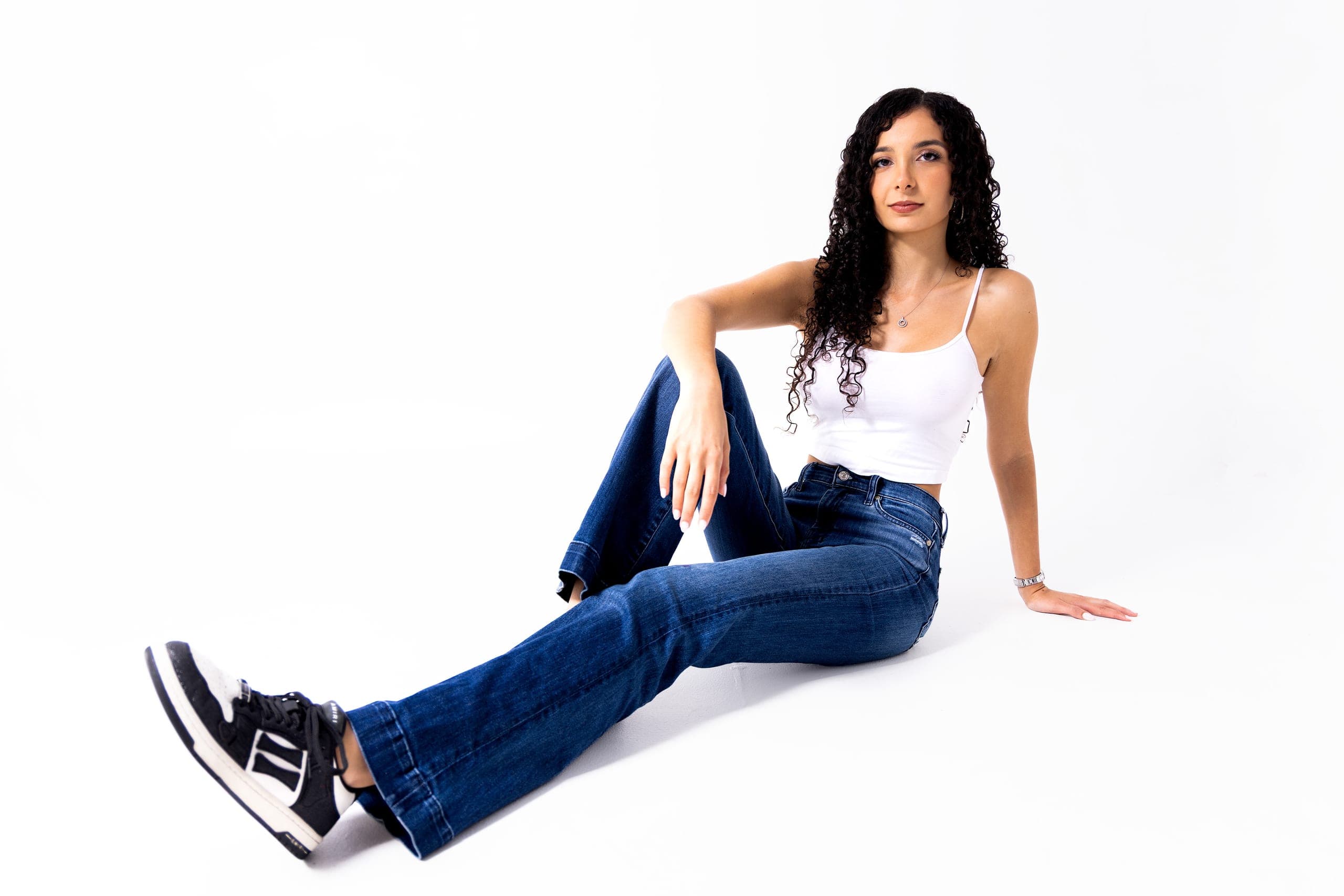 A studio portrait on a white seamless background, a woman seated on the floor in a white top, jeans and trainers, leaning back on one arm.